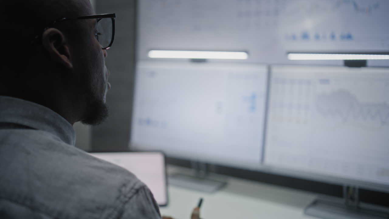 Financial Analyst Monitoring Real-Time Stocks, Exchange Market Charts on Computer with Multi-Monitor Workstation, Talking in Wireless Headphones. African American Businessman in Office. Vertical Shot.