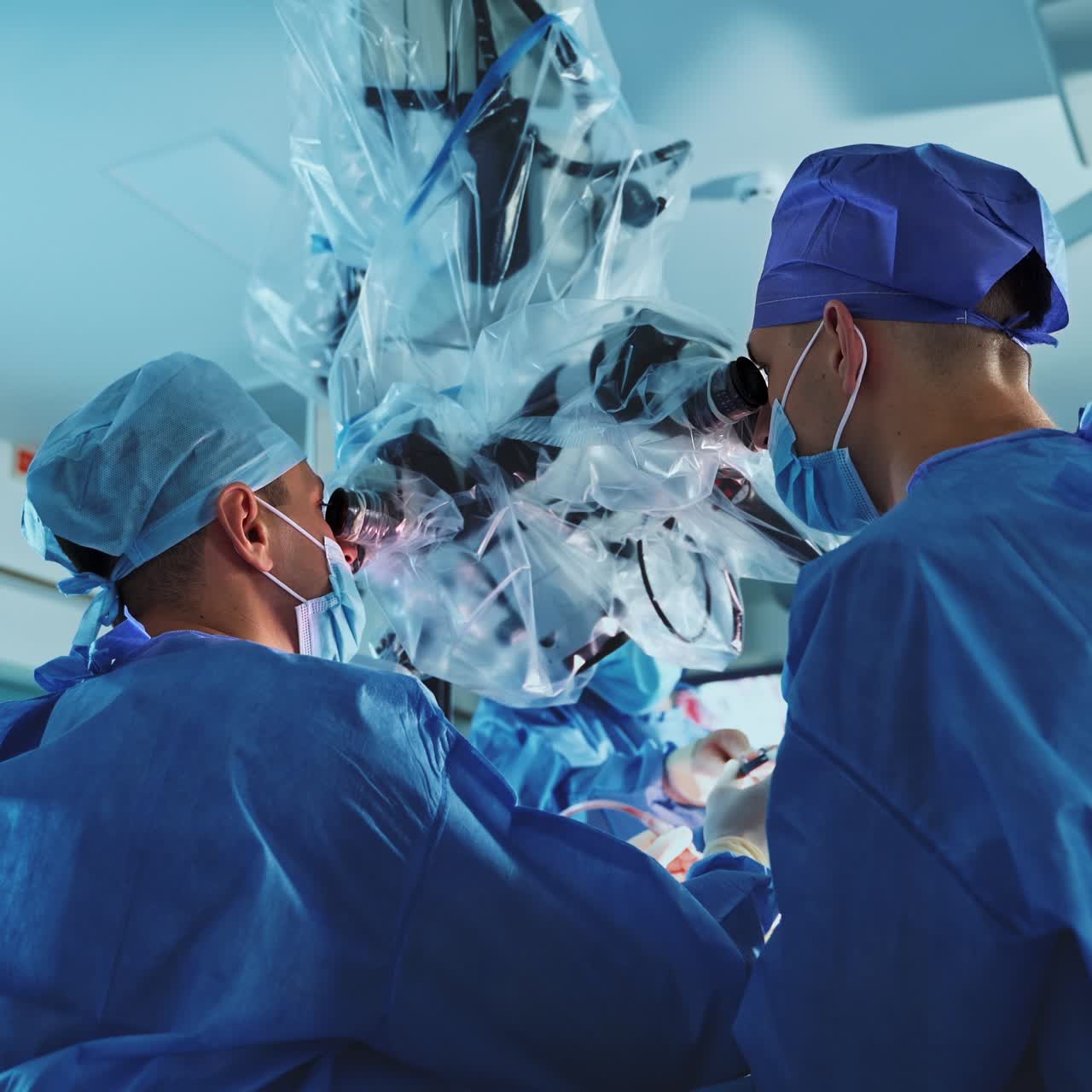 Two male doctors in blue uniforms, caps and masks conduct neurosurgery looking at powerful microscope. Low angle perspective on busy doctors