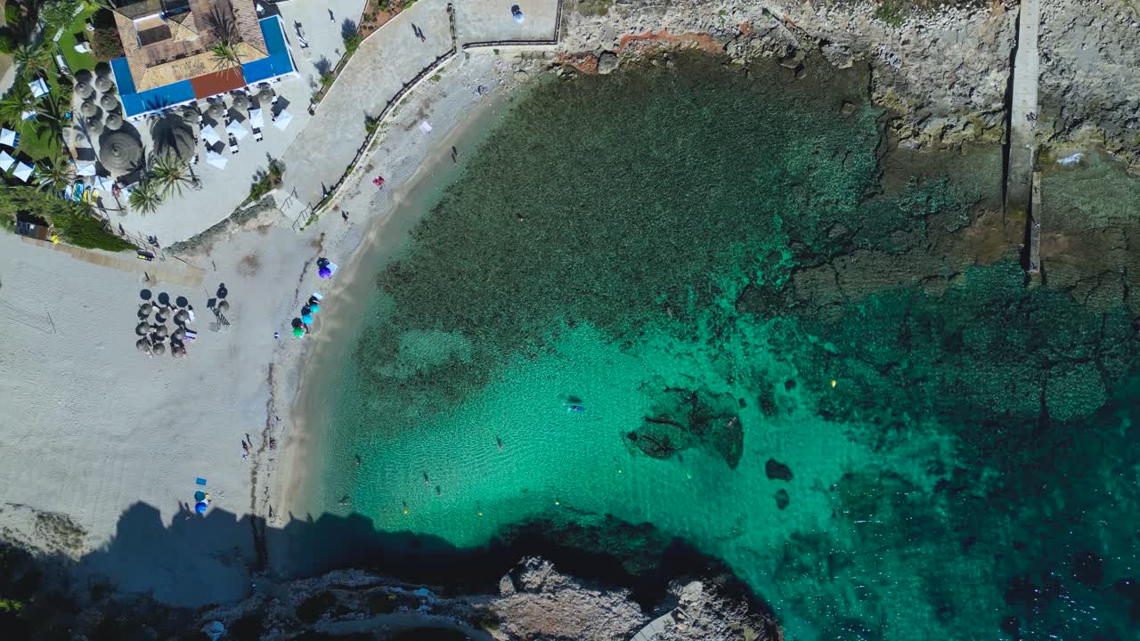 Stunning top-down aerial view of the picturesque Cala Murada cove and beach in Mallorca. The turquoise Mediterranean water, rocky coast, and surrounding hotels highlight the luxury travel destination