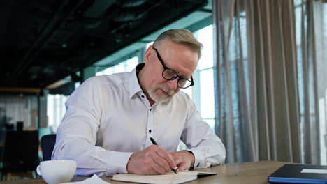 aged caucasian man sits at desk writing in his paper book. the pen stops writing and man blows on it and shakes it.