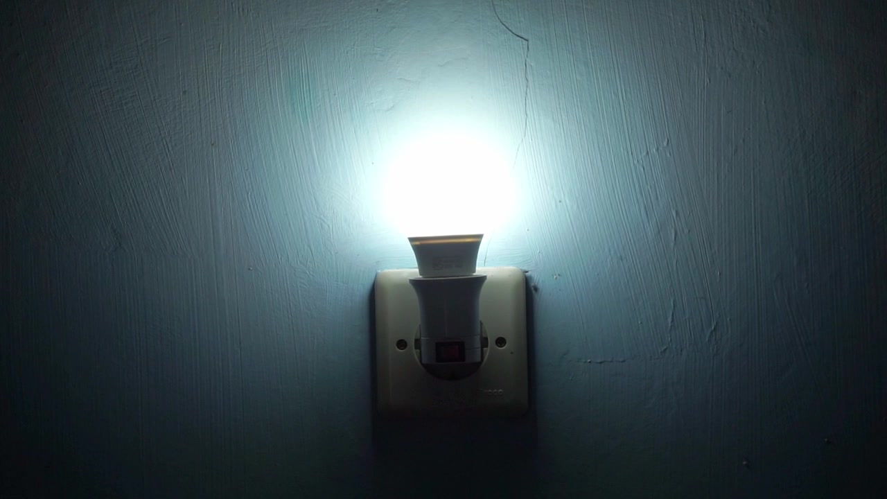 Hand turning on wall light switch, electricity and home lighting concept with glowing LED bulb in dark room interior.