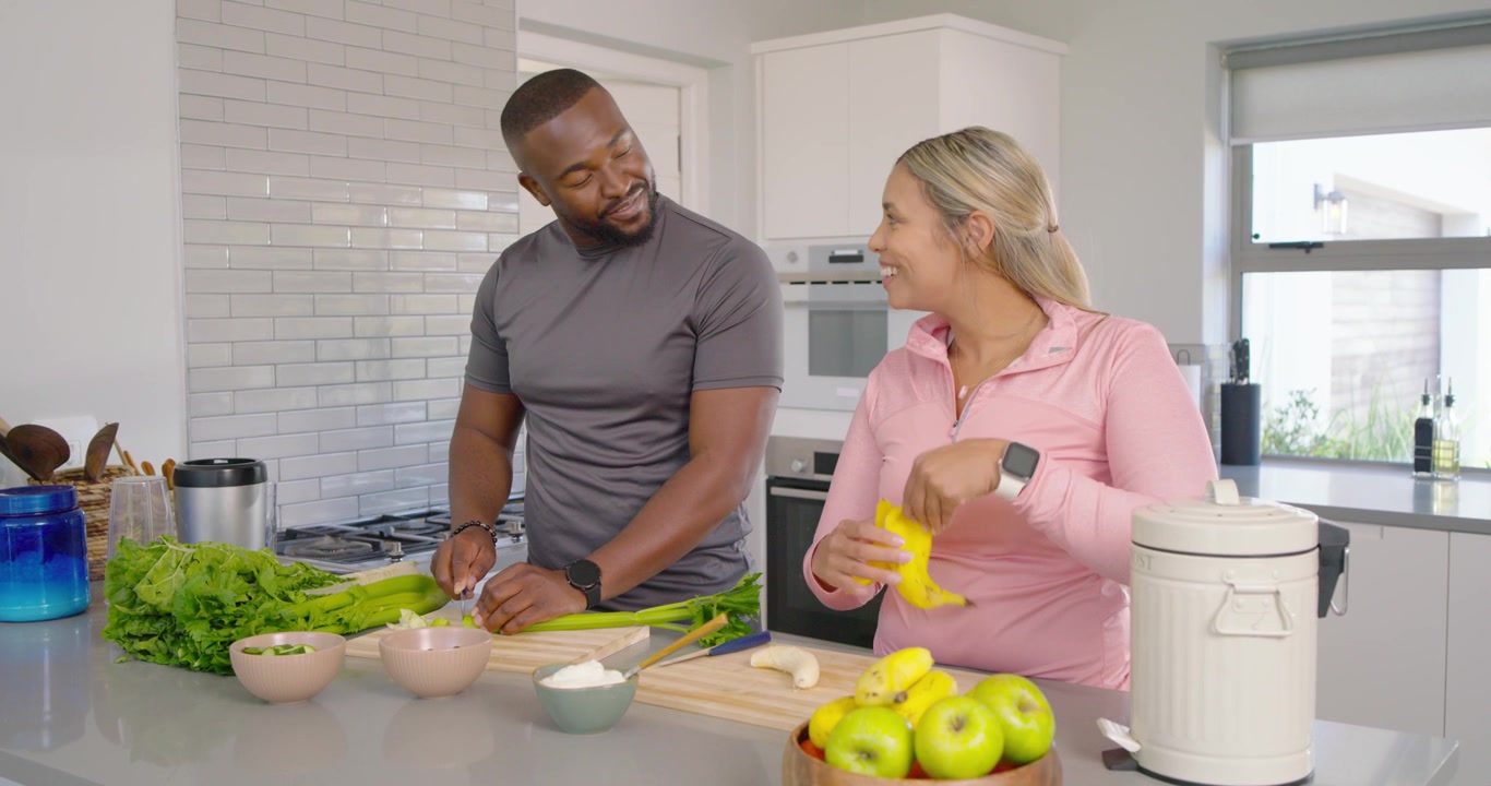 Casal preparando refeição na ilha da cozinha, o homem picando verduras em tábuas de madeira enquanto a mulher descasca uma banana.
