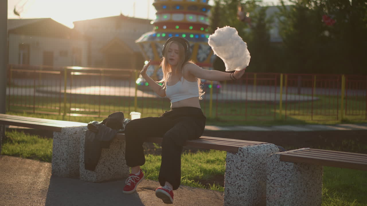 Parkta bankta dans eden, pamuk şekerini sallayan genç bir kadın, gün batımının fon ışığı ve lunapark ışıkları, gündelik sokak giyim tarzı ve kırmızı spor ayakkabılarıyla serbest ritimde coşkulu hareket, sokak sanatçısı enerjisi