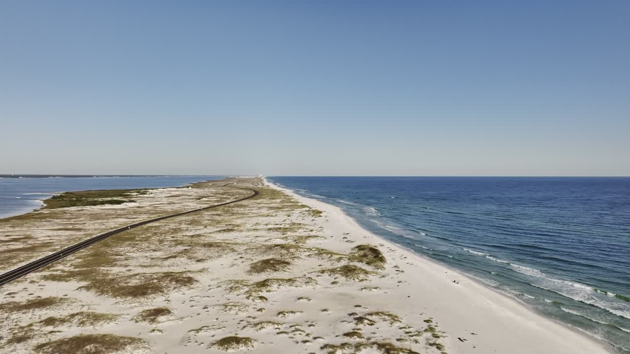 Wide elevation aerial drone shot of the Pensacola Beach tip road in Florida, capturing the scenic sunny coastal route and surrounding waters with clear blue sky and horizon in the background.