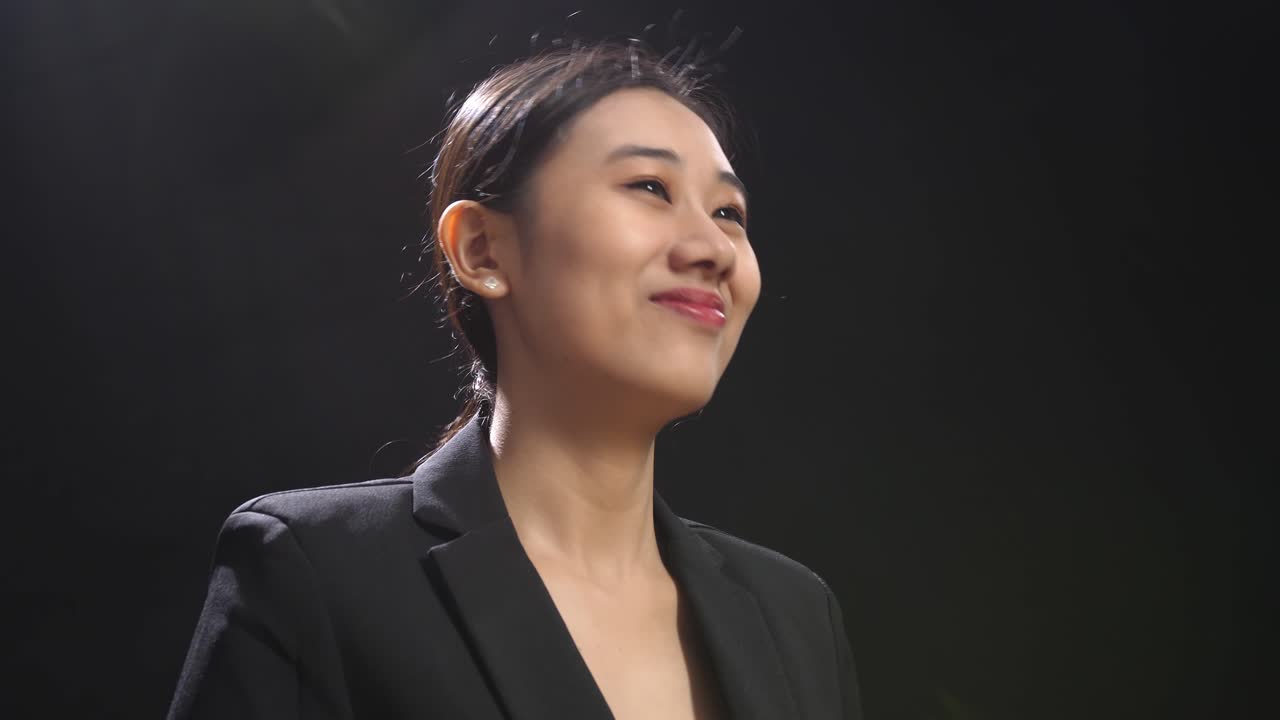 블랙 스크린 스튜디오에 서서 손을 치고 주위를 둘러보는 미소 짓는 아시아 스피커 여성