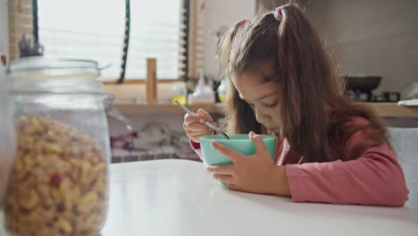 A-little-brunette-girl-in-a-pink-dress-sits-at-the-table-and-eats-cereal-with-milk-and-from-a-blue-plate-in-a-modern-apartment-in-the-morning.-Little-brunette-girl-has-breakfast-sitting-at-the-table-in-a-modern-apartment