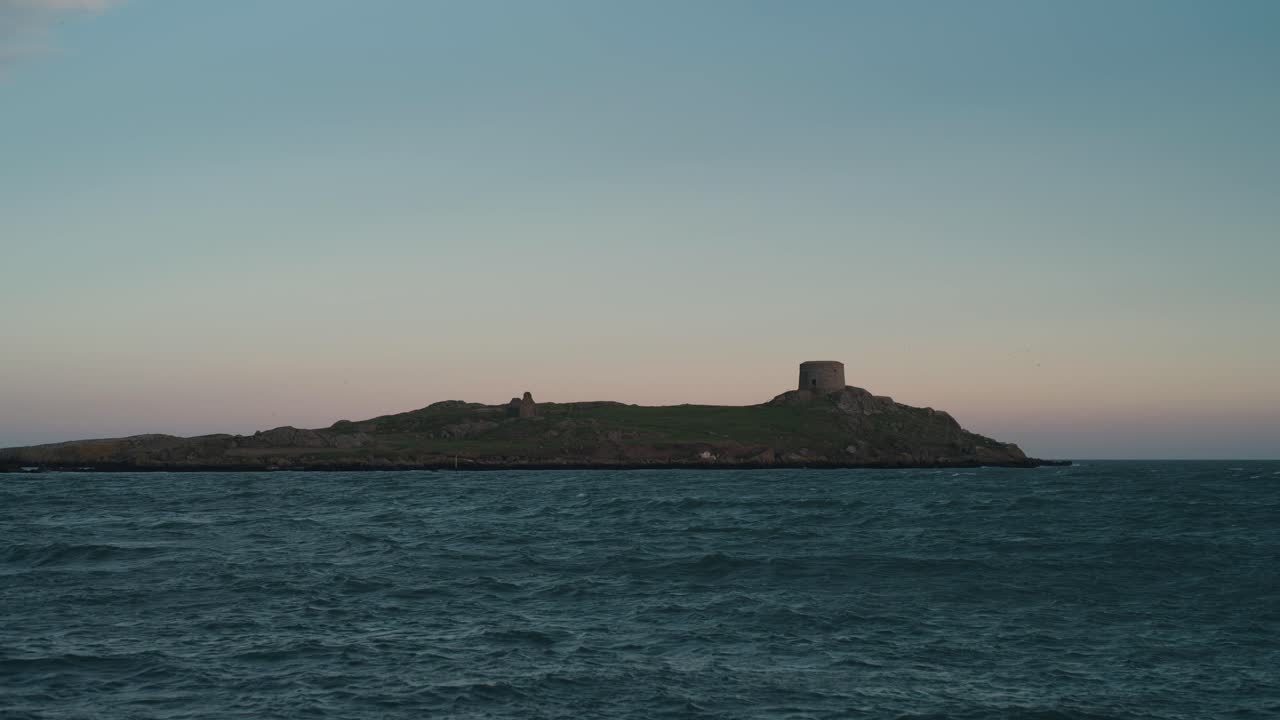 A locked-off shot of Colliemore Harbour in Dublin, Ireland. The stone pier leads toward Dalkey Island as gentle waves roll under a soft pastel sunset sky.