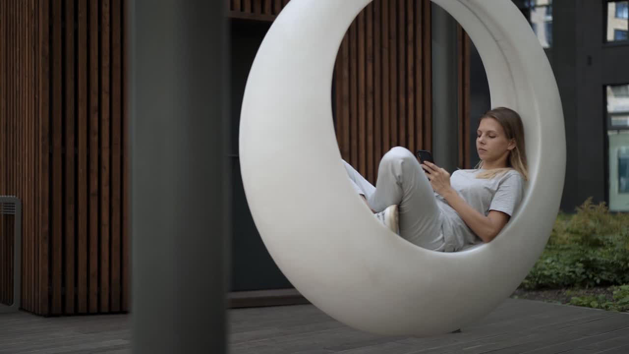 Young woman sitting in white futuristic swing in the yard, gimbal shot