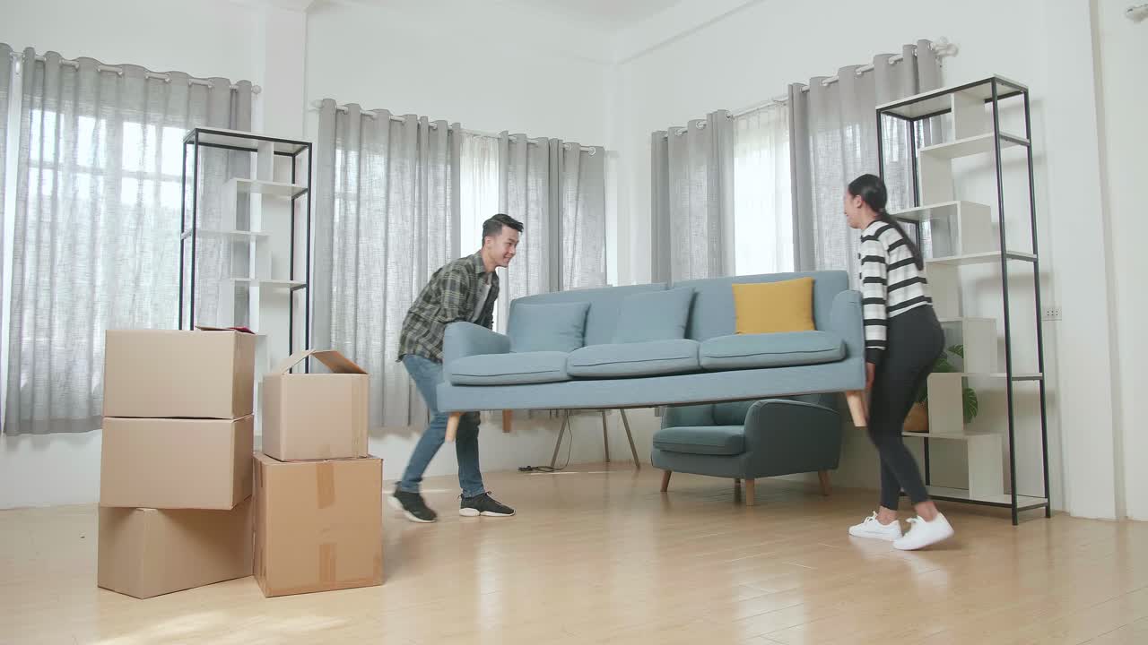 행복한 젊은 아시아 부부는 휴식을 취하기 전에 새로운 소파를 새로운 집으로 옮기고 있습니다.