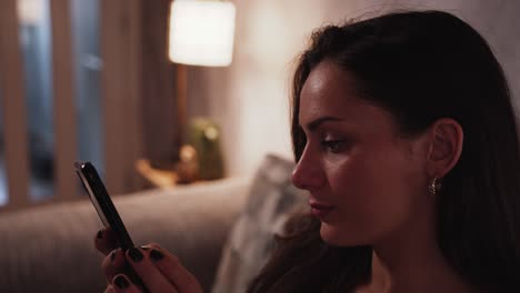 Young-woman-using-social-media-on-a-smart-phone-on-the-sofa-in-a-cosy-home