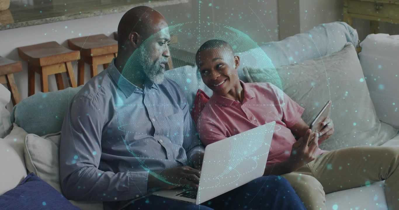 Typing gray-shirt man leaning toward red-shirt teen showing tablet on home couch, laptop teal-globe