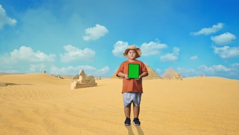 asian boy with a hat smiling and showing green screen tablet. boy researcher examines something while traveling in giza pyramid, travel tourism adventure concept, full body