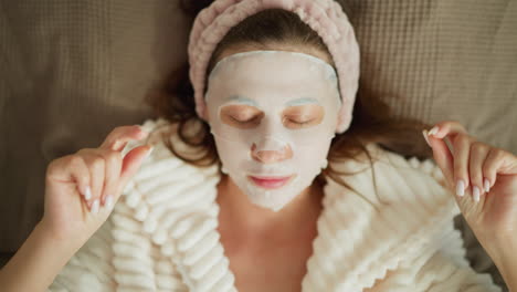 lady puts relaxing mask enjoying pleasant leisure time. brunette woman with facial mask lies on soft surface listening to pleasant music in room