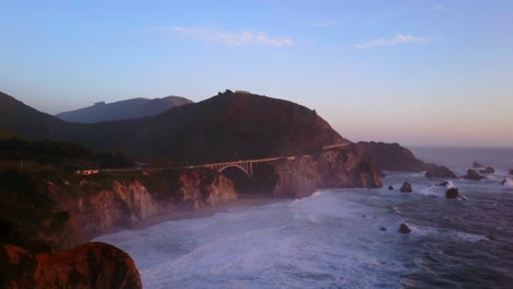 bixby creek bridge big sur california vuelo aéreo cinematográfico de drones océano pacífico ni cal invierno verano gran oleada se hincha chocando contra la costa tarde hora de oro puesta de sol hacia arriba movimiento lento