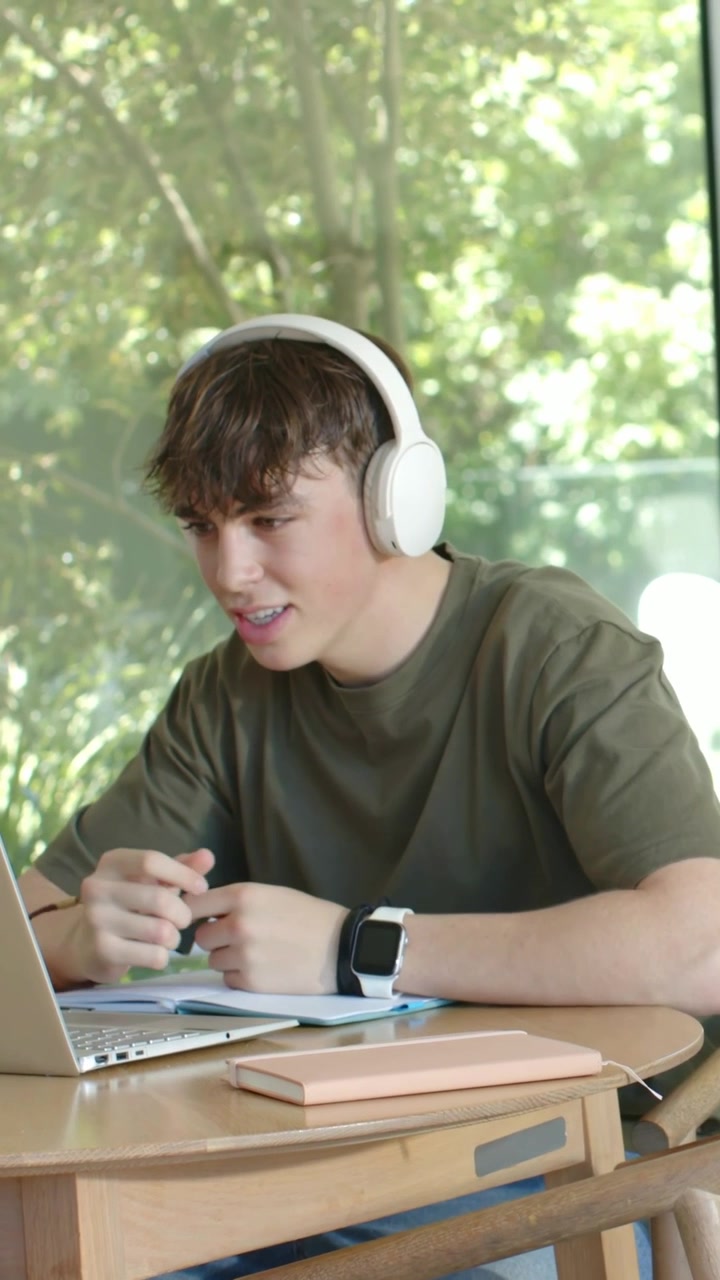 Male student wearing headphones joining online lesson on laptop, leaning and explaining notes