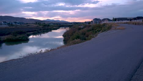 person riding a bike along a trail as a stunning sunset reflects on the river - riding away from static camera