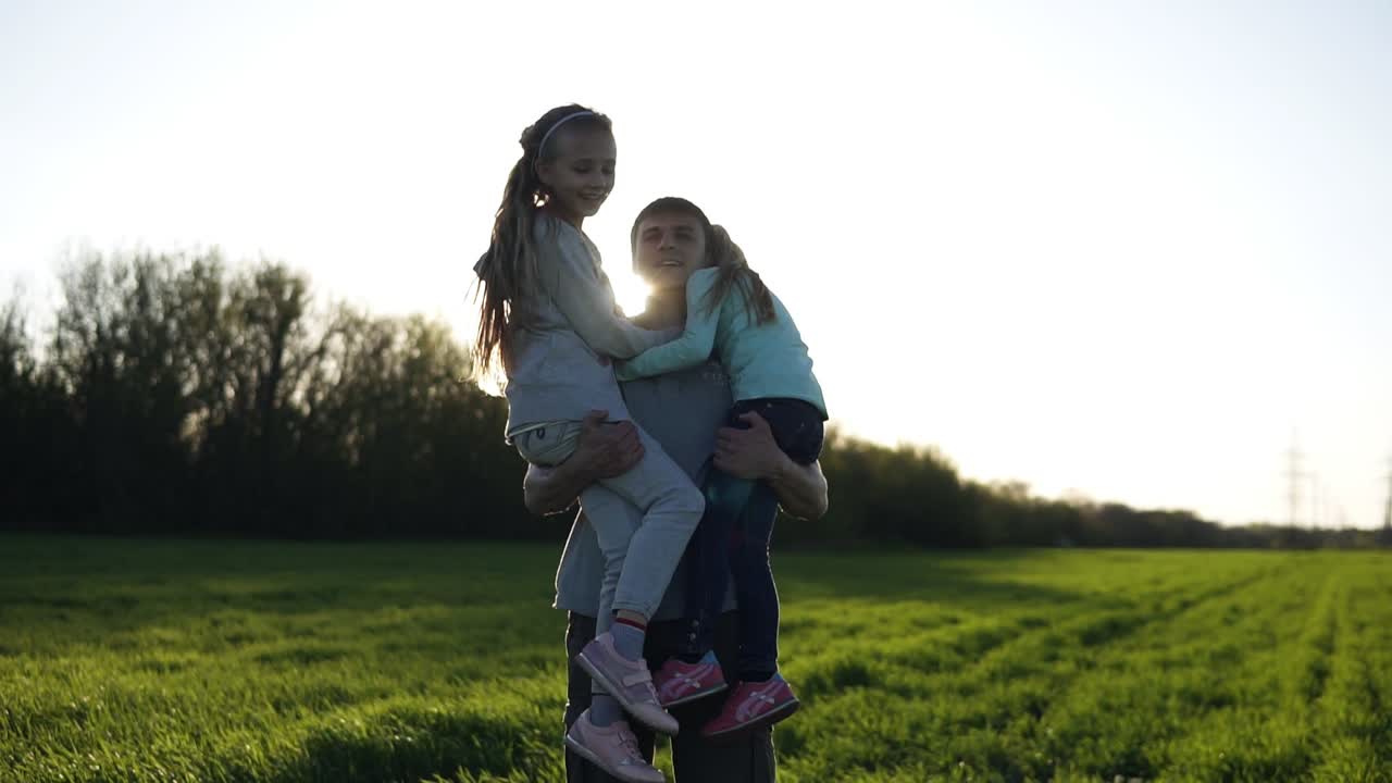 Proud father of two girls in nature. He carries them in his arms, the sun is shining, the youngest girl sends air kisses. Beautiful field with green grass. Slow motion