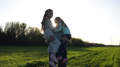 orgulloso padre de dos niñas en la naturaleza. las lleva en sus brazos, el sol está brillando, la niña más joven envía besos al aire. hermoso campo con hierba verde. cámara lenta