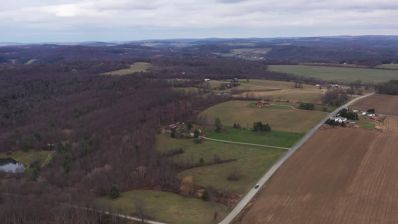 Penerbangan pandangan drone udara di atas rumah, lahan pertanian, hutan, ladang hijau, dan jalan, melacak pergerakan mobil pada hari musim gugur di pedesaan Roma, Pennsylvania.