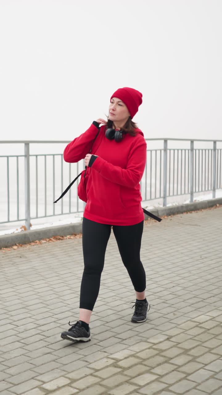 Freelancer in red winter jacket adjusts bag over shoulder while walking along interlocked pathway near iron railing with dry scattered foliage in foggy atmosphere, distant car visible below bridge