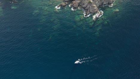 Toma-Aérea-Del-Pequeño-Bote-Blanco-Navegando-En-Las-Aguas-Esmeralda-Cerca-De-Porto-Cervo,-Cerdeña,-Italia