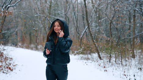 long-haired brunette girl walks by the park speaking on the phone. smiling lady holds a paper cup with coffee. winter nature at backdrop.