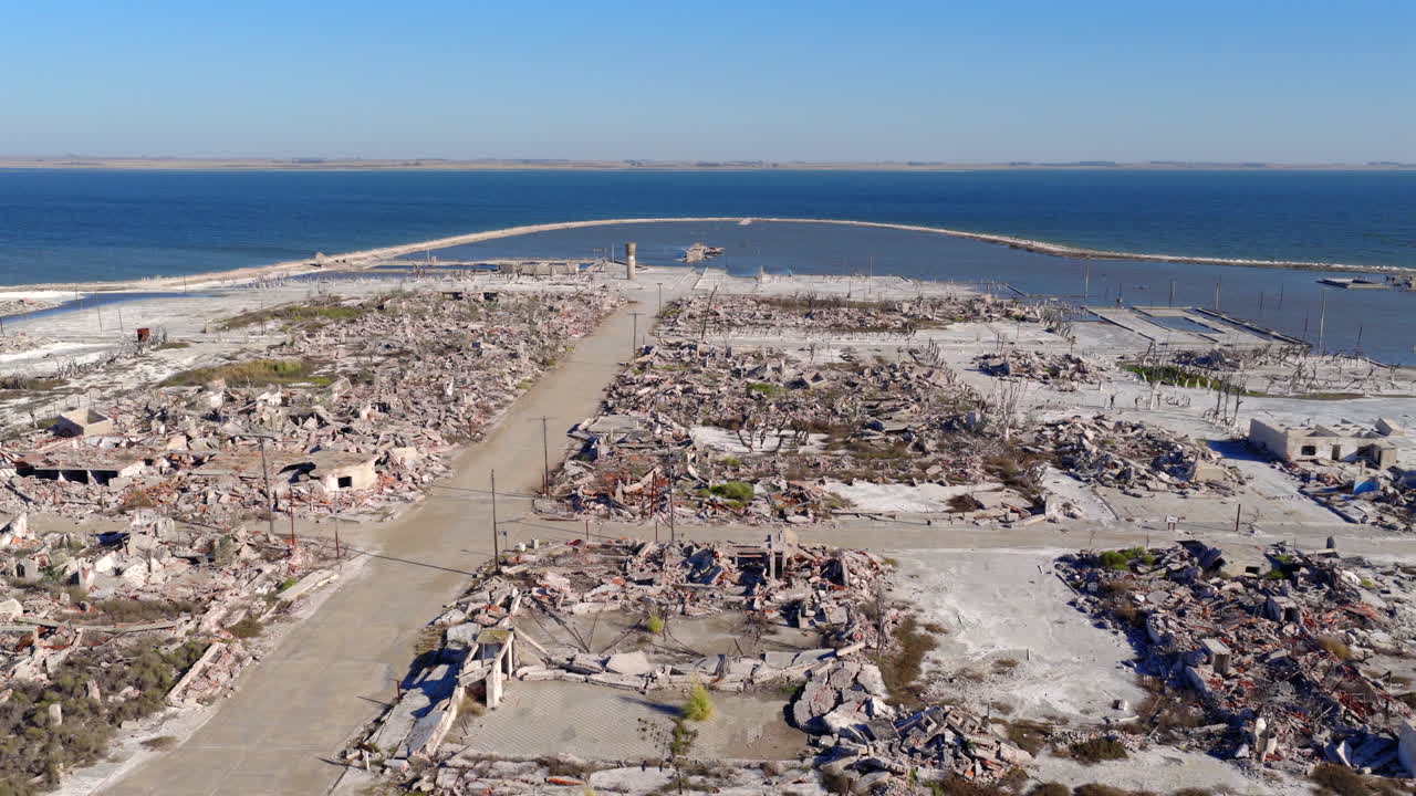 Drone camera frames the main avenue of Villa Epecuén, shooting over rows of destroyed and salt-covered buildings, converging toward the old pier and the expansive blue waters of the lake