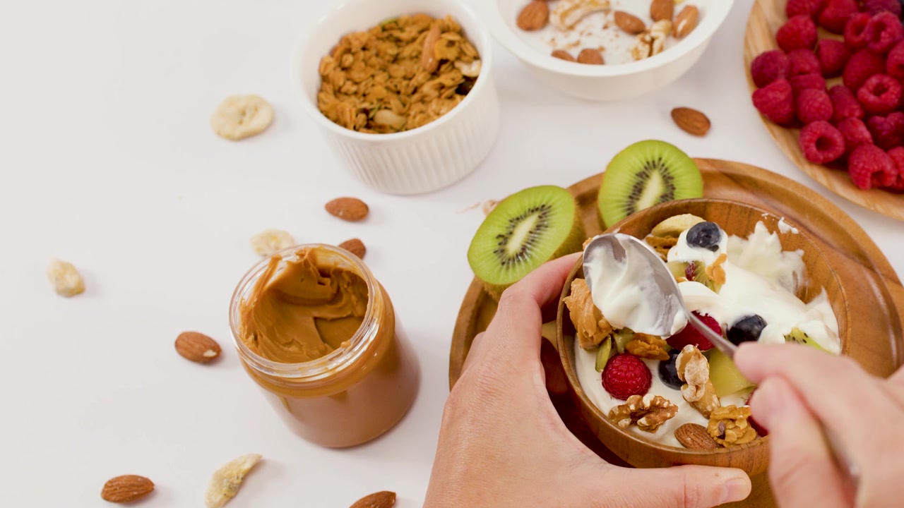 Overhead shot of hands scooping yogurt, berries, and kiwi from a wooden breakfast bowl.