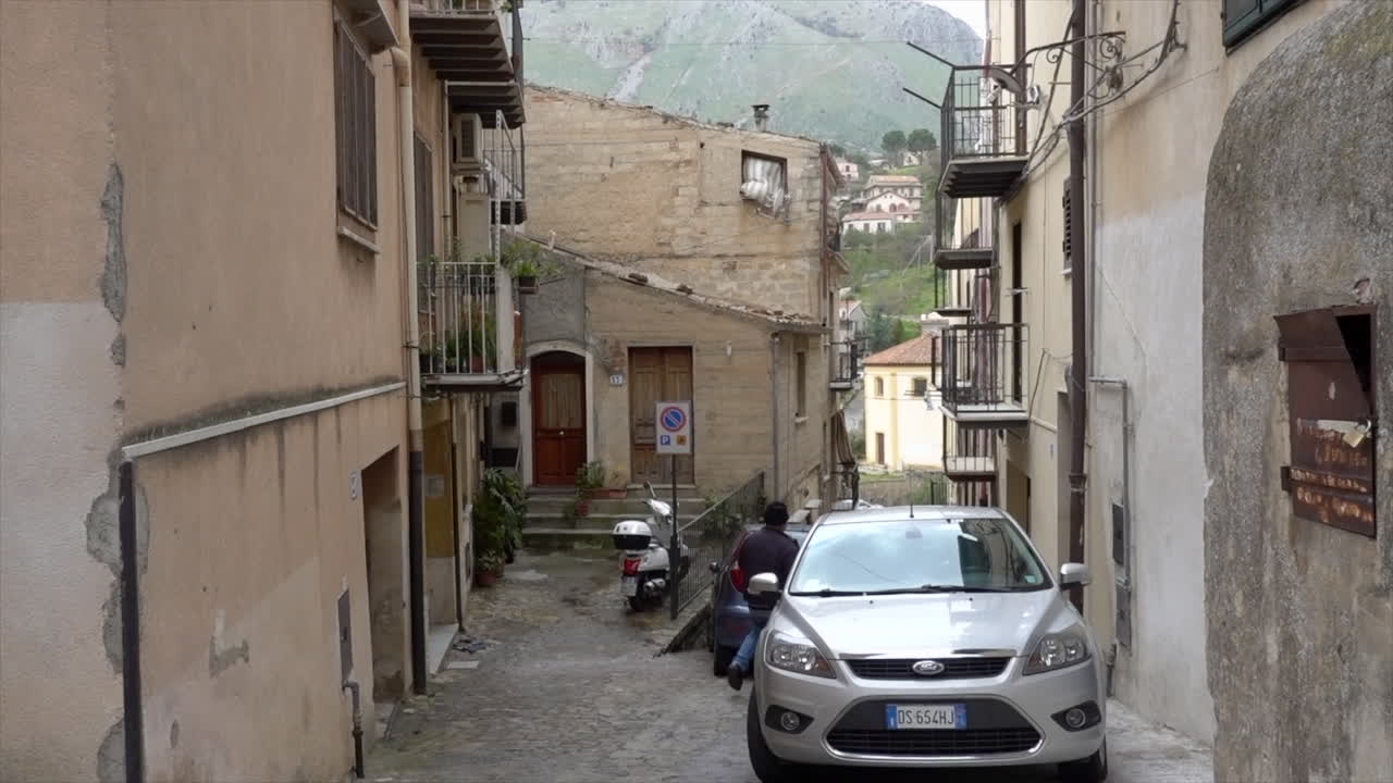 Street view of the town on Castelbuono. A narrow street with cars parked on the side and a person walking