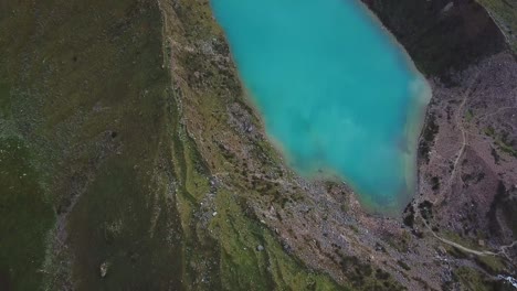 Aerial,-tilt-up,-drone-shot-overlooking-a-crystal-clear,-emerald-water-of-lake-Humantay,-Andes-mountains,-cloudy-day,-in-Peru,-South-America