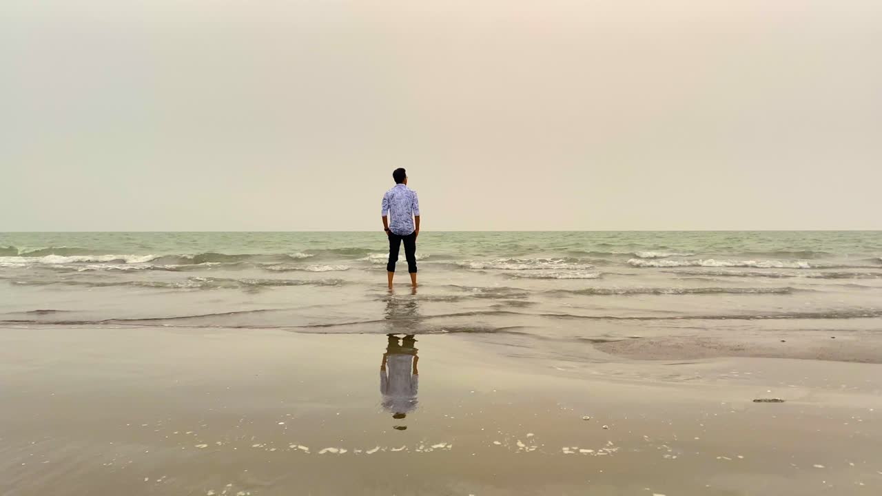 A lone man stands at the quiet shoreline, gazing toward the horizon as soft waves reflect his silhouette, capturing a calm yet thoughtful moment of solitude on a tranquil beach