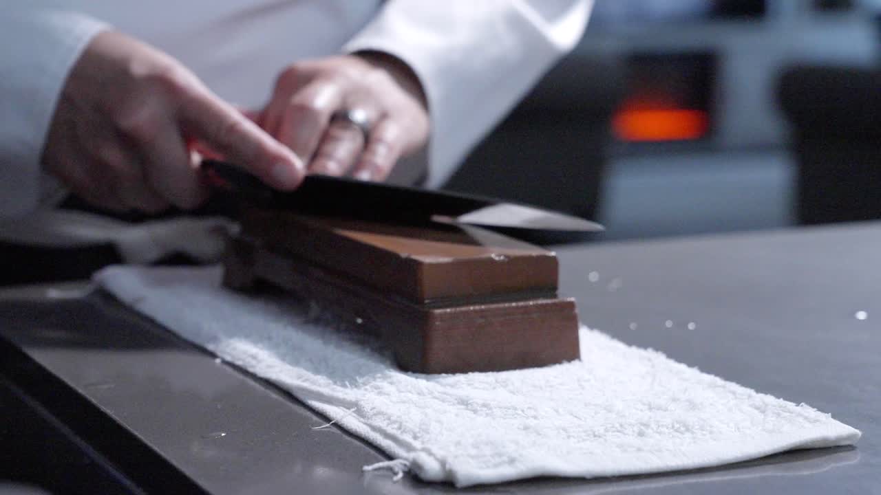 Inquadratura statica laterale sinistra di uno chef che affila il suo coltello, evidenziando la tecnica abile e l'attenzione alla manutenzione del coltello. Perfetto per tutorial di cucina e dimostrazioni culinarie.