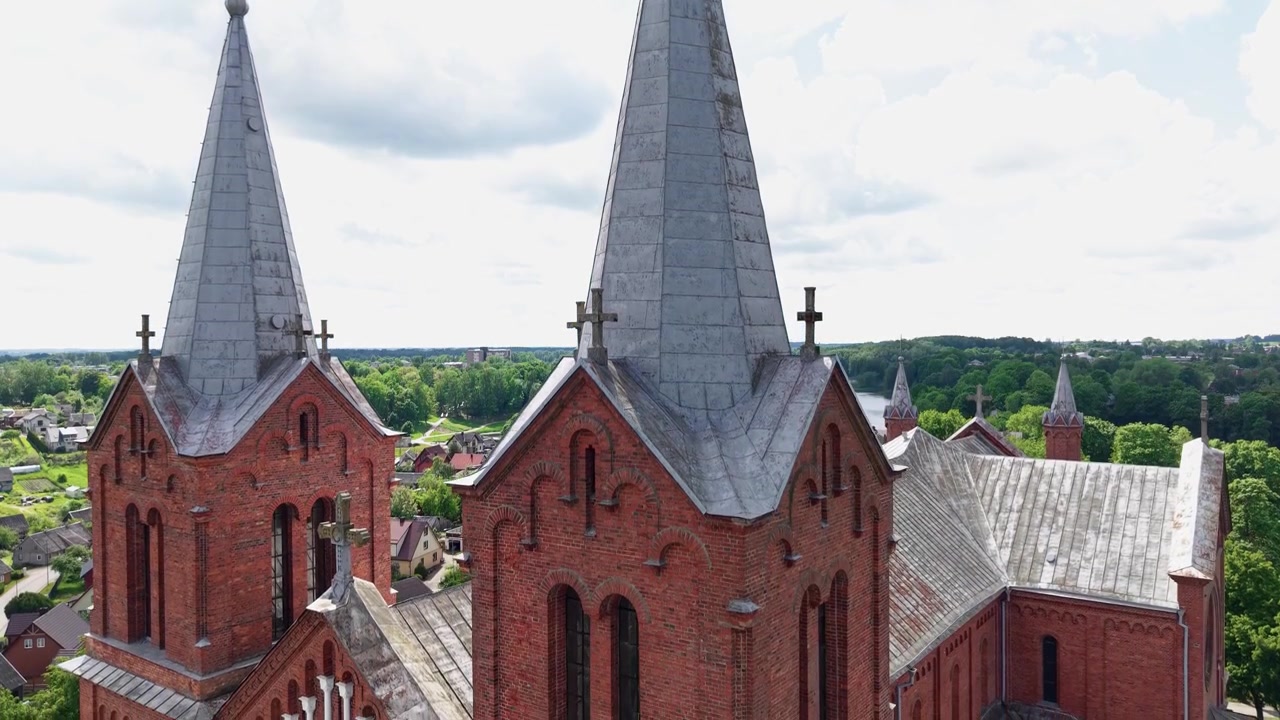 Imponerande drönarfilm visar upp de nygotiska spirorna från Johannes Döparens kyrka i Plungė, Litauen, mot frodig grönska och en glittrande sjö i dynamiskt dagsljus