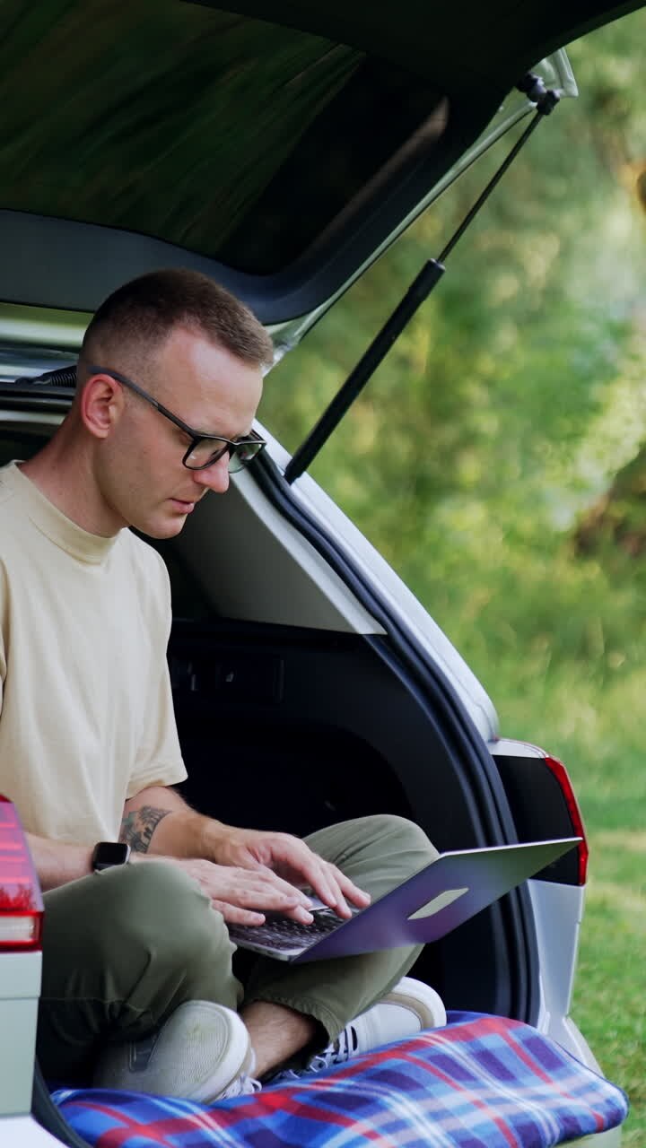 Uomo freelance lavora al suo computer portatile. Imprenditore lavora all'aperto seduto nel bagagliaio della sua auto. Video verticale