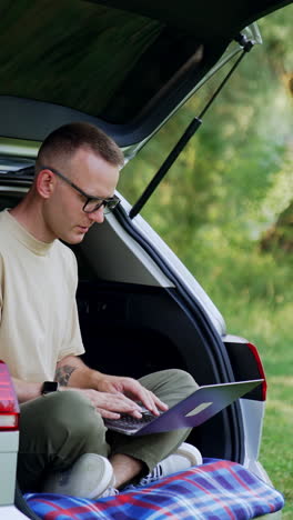 freelancing man works on his laptop. entrepreneur works outdoors sitting in his car trunk. vertical video