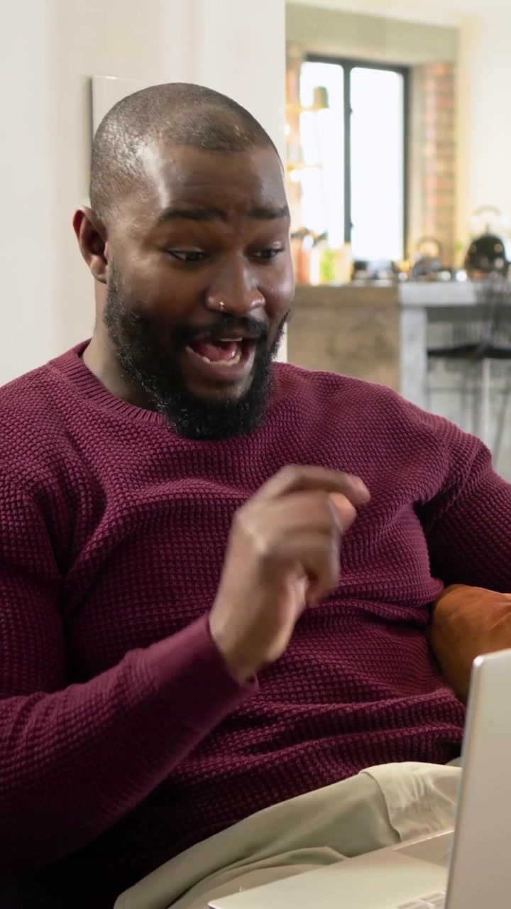 Video verticale: Uomo sorridente con un maglione color vino che guarda il laptop, digita sul divano con un cuscino arancione