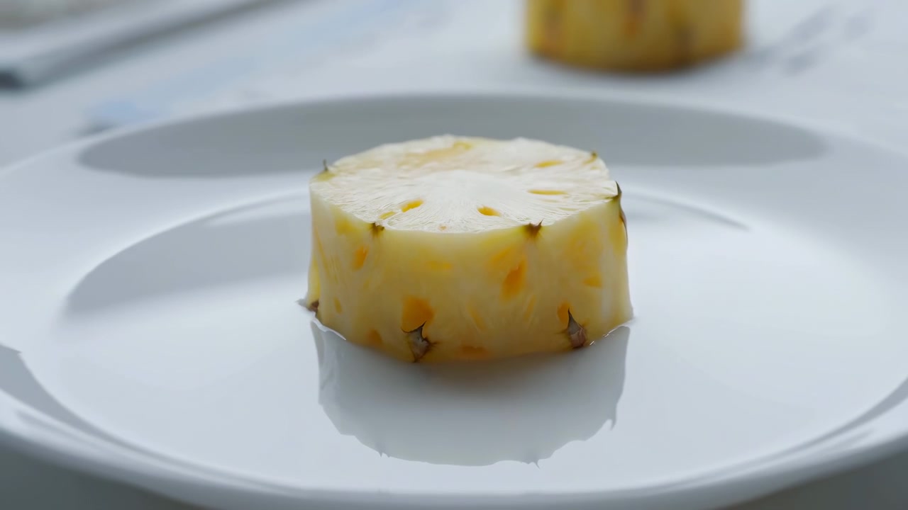 Una singola fetta di ananas centrata su un piatto di ceramica bianco lucido, che mostra la buccia tagliata, polpa fibrosa giallo pallido, piccoli occhi scuri, piccole protuberanze marroni, luci soffuse, riflesso e studio minimale.