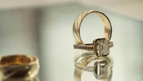 two gold rings, one with a diamond, are displayed on a reflective surface, one ring spinning