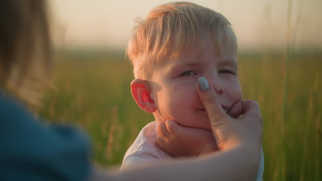 a close-up shot capturing the tender moment as a mother s hand wipes her young son's face, focusing on his eyes, nose, and mouth. the boy, with a serious expression, is in a grassy field during sunset