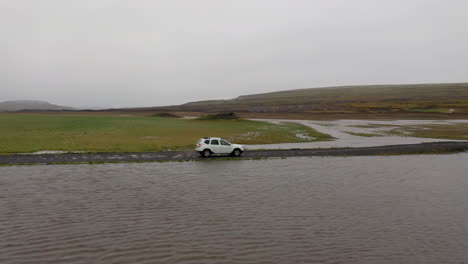 aerial: side follow of white car passing through a shallow river