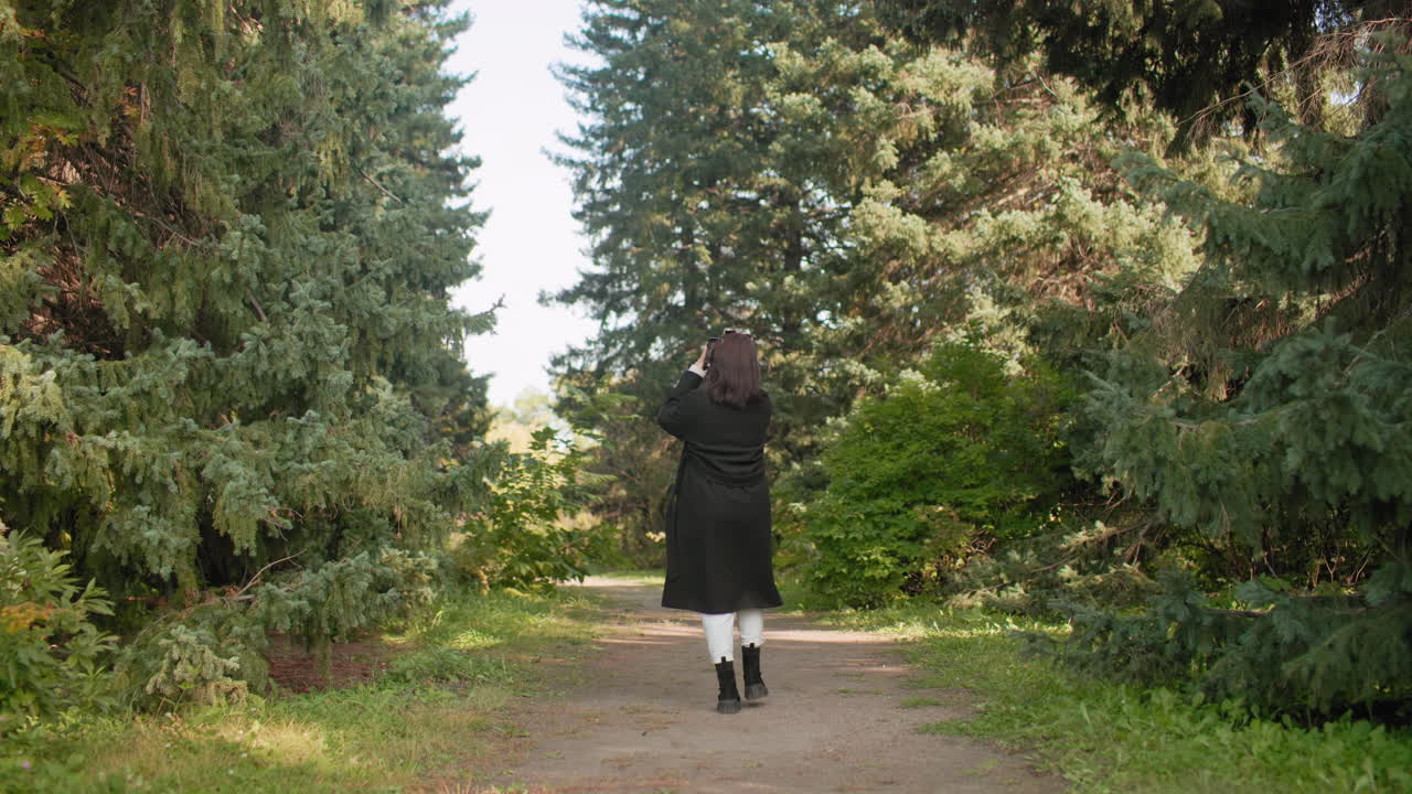 숲길에서 핸드폰을 들고 사진을 찍으려고 폰을 든 채 걷는 뒷모습, 키 큰 소나무 아래 긴 코트, 머리에 선글라스, 조용한 가을의 초록, 산책하며 풍경을 담는 모습이에요.