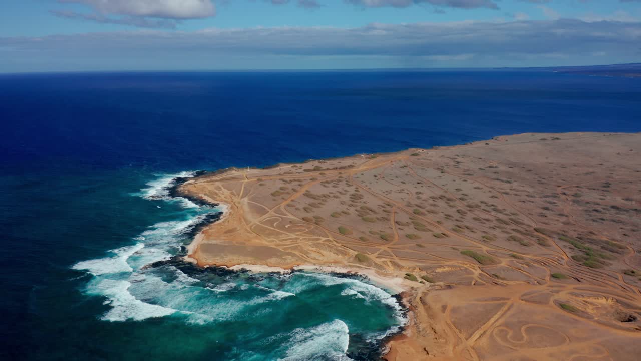 A drone glides above a barren coastal headland scored with off-road tracks, where turquoise waves batter dark cliffs and foam curls into coves, evoking isolation and windswept energy