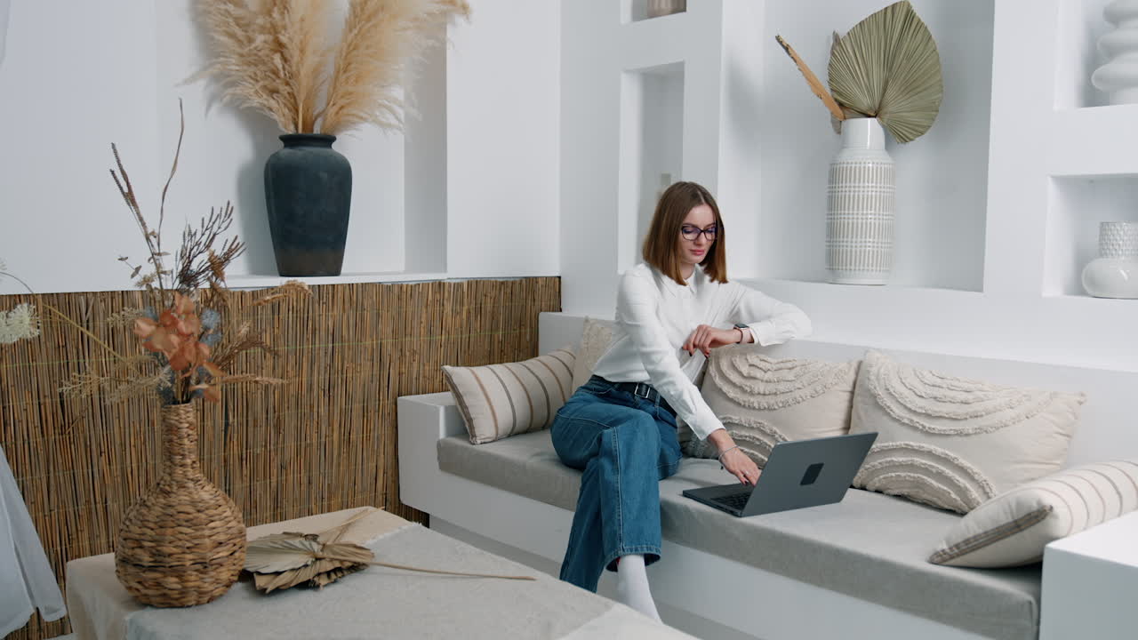 Relaxed positive Caucasian woman sitting in the room on the sofa. Woman looks at her laptop and presses some keys on keyboard.
