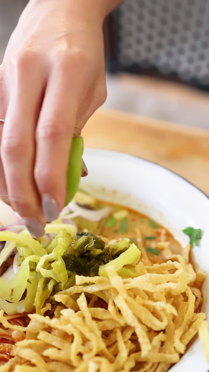 Una mano spreme lime fresco su una ciotola di noodles al curry di manzo con guarnizioni croccanti, in un ristorante thailandese al coperto ben illuminato, ripreso in primo piano.