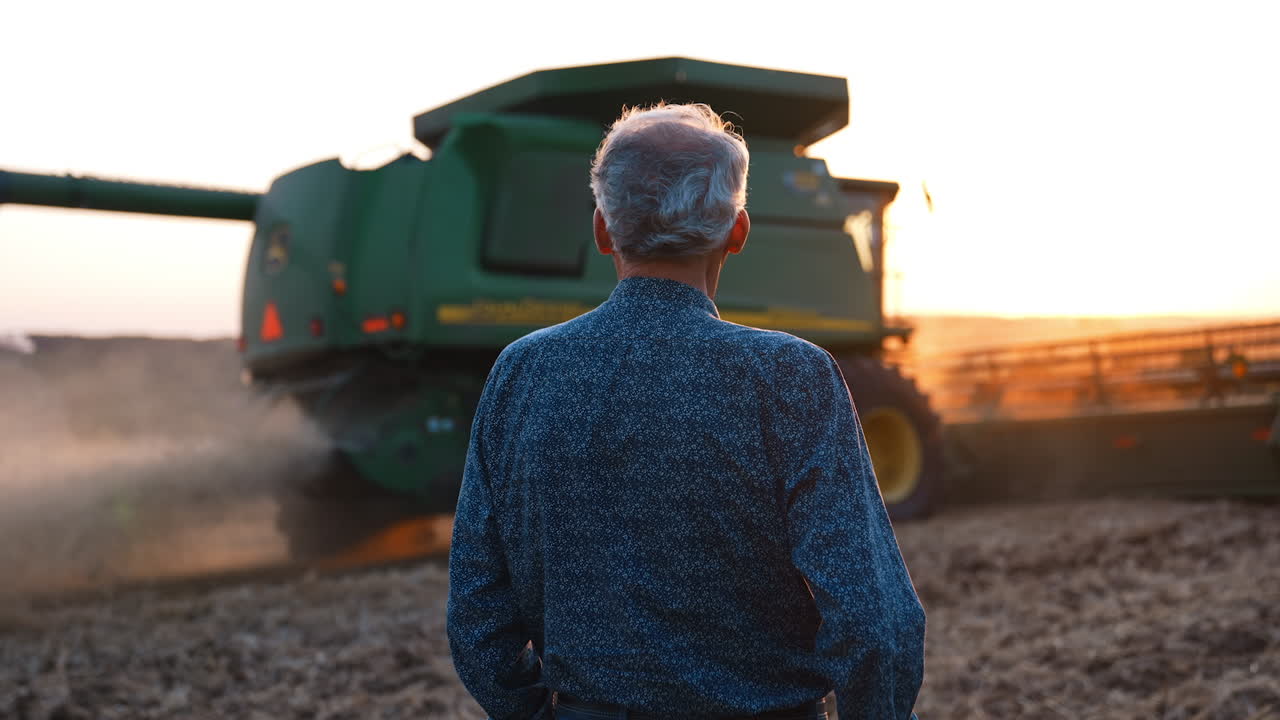 Farmer observing sunset over fields. A farmer stands in a field, watching a combine harvester work as the sun sets, creating a serene atmosphere