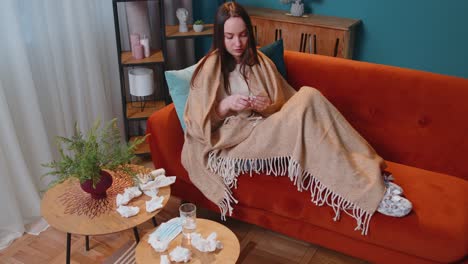 unhealthy sick young woman sitting sofa in warm plaid taking medicine pills tablets drinking water