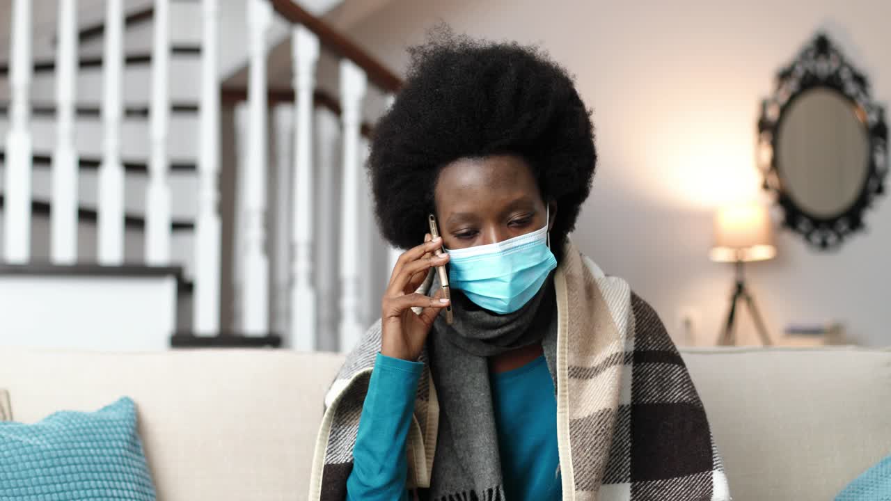 Close up portrait of sick woman wearing a scarf, sitting in room and calling on cellphone while coughing