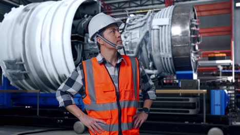 asian male engineer wearing safety helmet looking around while standing with arms akimbo with airplane engine maintenance conducted, analytics checking the turbine