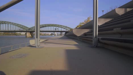 low dolly forward shot between handrails of stairs in a sitting area next to a river with a steel arched bridge in the background on a sunny clear day, slow motion