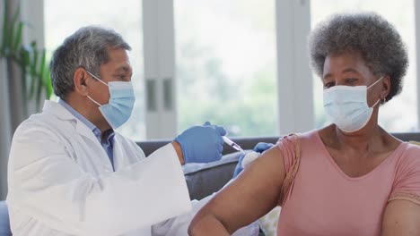 male doctor wearing face mask vaccinating african american senior woman at home